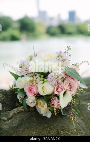Pastel roses and lush greenery create an elegant bouquet displayed on a rock by a calm lake under clear skies. Stock Photo