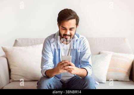Happy arab man using smartphone at home, messaging with friends Stock Photo