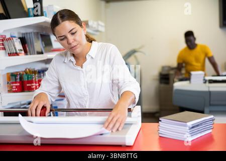 Focused young female typographer trimming the printed work on the cutter in the typography Stock Photo
