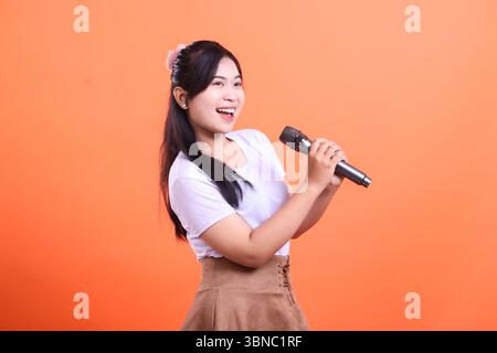 A cheerful young woman holding a microphone, performing or singing with a big smile on her face, enjoying her hobby, isolated on orange background Stock Photo