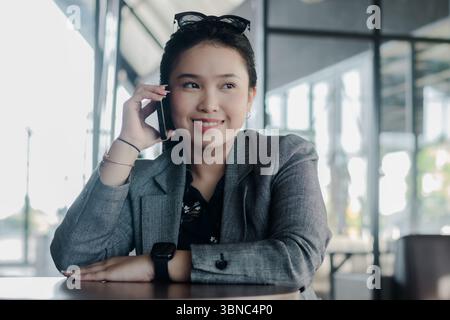 A young Asian businesswoman is engaged in a phone conversation, radiating happiness. She is smartly dressed in casual attire, reflecting a modern and Stock Photo