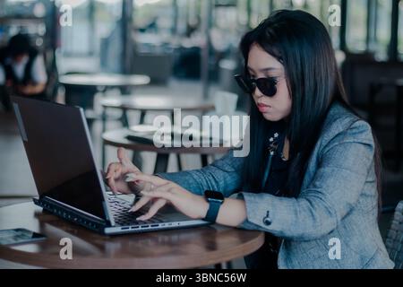 A focused businesswoman in sunglasses working on her laptop in a stylish caf�nvironment. She is seen actively typing, suggesting a professional and pr Stock Photo