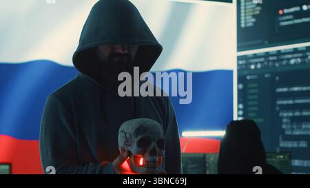 Conceptual image of a hooded russian hacker creating hazardous alerts, holds a skull and warning of death threats. Russian federation works on cybercrime and violence, panic and torment. Camera A. Stock Photo