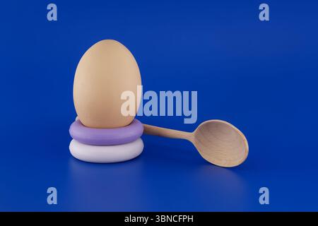 A brown egg rests on a stack of soap rings, complemented by a wooden spoon against a vibrant blue backdrop. Stock Photo