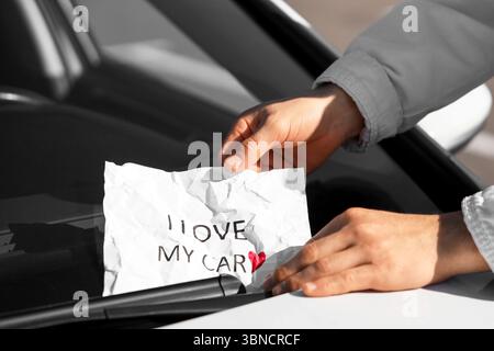 Young man putting sheet of paper with heart and text I LOVE MY CAR on windshield, closeup Stock Photo