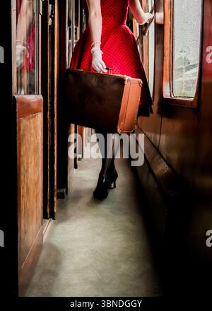 A woman in a red polka dot dress, white gloves, and black heels walks down the aisle of a vintage train car, carrying a brown suitcase. Stock Photo
