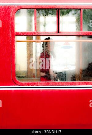 A woman in a red dress is seen through a window of a red vehicle, possibly a bus or train, with raindrops on the glass. Stock Photo