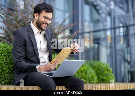 A smiling businessman is reading a document while working on his laptop outdoors, in front of a modern glass building. Stock Photo