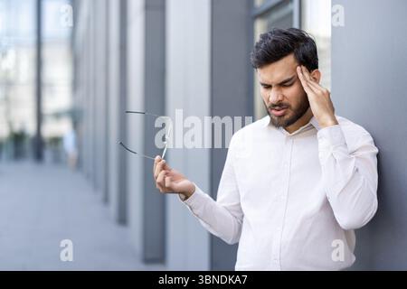A man experiencing a headache, holding glasses, and leaning against a wall outside of a building. Stock Photo