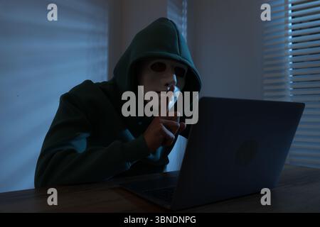 Anonymity. Masked man with laptop showing hush gesture at table in dark Stock Photo