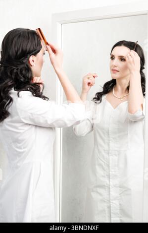 A woman, wearing a white coat, stands before a mirror, carefully combing her dark hair. The reflection shows her focused expression. Stock Photo