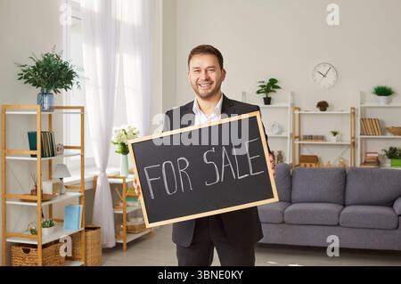 Portrait of cheerful young man realtor holding a sign for sale standing in living room. Stock Photo