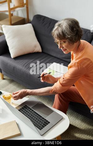 Elderly woman in comfortable attire engaged in an online task while sipping tea. Stock Photo