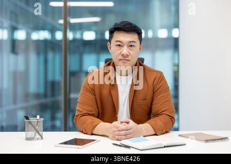 A focused Asian man sits at a desk, looking directly at the camera in a modern office setting, wearing a button-down shirt. Stock Photo