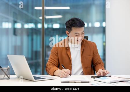 An asian man is working at his desk, smiling while using a calculator and writing on a document in an office. Stock Photo