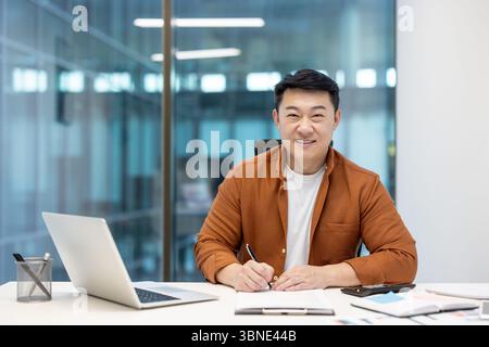A smiling asian businessman working at his desk, writing and looking at the camera. His office is modern. Stock Photo