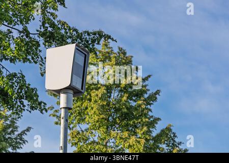 A modern speed camera stands tall amidst green trees against a bright blue sky in the Netherlands, symbolizing traffic enforcement and urban safety. Stock Photo