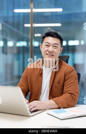 An Asian man smiling while working on a laptop in a modern office environment. Stock Photo