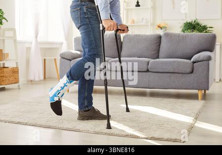 Young man with broken leg after accident using crutches standing at home with plastered leg. Stock Photo