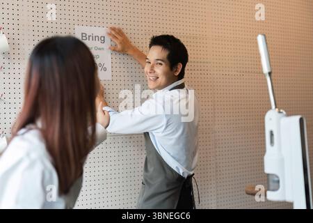 Eco-Friendly Promotion and cafe Community. A barista playfully engaging with her colleague while putting up a sign to encourage customers to bring Stock Photo