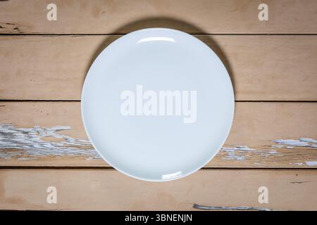 Close-up top-down view of a white porcelain plate centered on a distressed beige wooden table with cracked, peeling paint and visible texture. Stock Photo