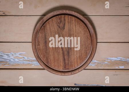 Close-up top-down view of a round wooden cutting board with visible knife marks on a distressed beige wood table with peeling paint. Stock Photo