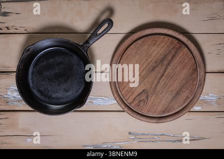Top-down view of a small cast iron skillet next to a round wooden cutting board with knife marks on a beige distressed wooden table. Stock Photo