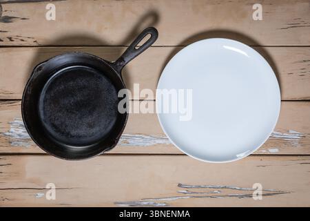 Top-down view of a cast iron frying pan and a white porcelain plate placed side by side on a distressed beige wooden table. Stock Photo