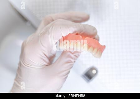 A gloved hand holds an upper denture with visible wear, isolated in bright light on a neutral background, showing signs of realistic use. Stock Photo