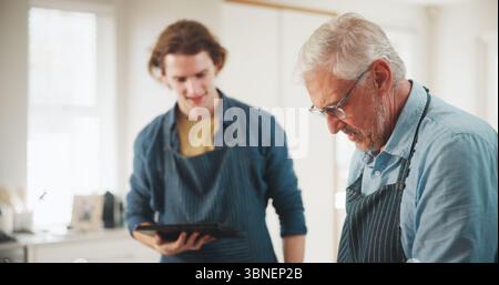 Mature man, chef or cooking with tablet in kitchen for culinary skill or hospitality in home. Male people, mentor and helping assistant with Stock Photo