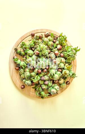 Vertical top view of organic hazelnuts in green husks and shells on a wooden plate. Minimalist seasonal food layout on pastel yellow background with c Stock Photo