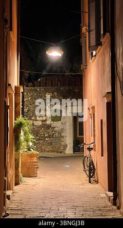 A serene nighttime alley scene features a vintage bicycle parked against a wall with an old lantern illuminating the path. Rustic stone and aged brick Stock Photo