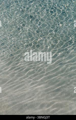 Gentle waves create patterns on the clear water over the soft sand, inviting relaxation under the warm sun. Stock Photo