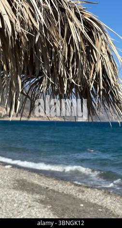 Close-up of a straw umbrella providing shade on a sunny beach day, with gentle waves rolling onto the shore in the background. The image captures a pe Stock Photo