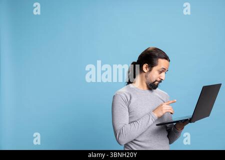 Cheerful and focused young person using his pc for online tasks, embodying the concept of digital connectivity. Man with long hair browsing the web for modern work or internet communication. Stock Photo