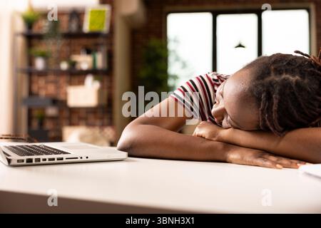 African american female freelancer naps on desk beside laptop in home office, physically drained from working under tight deadline. Woman entrepreneur peacefully sleeping after completing tasks. Stock Photo