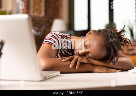 Black female entrepreneur lies on desk in home office, taking quick nap after demanding morning of work. Tired remote worker rests near laptop, overwhelmed by exhaustion from completing tight deadlines Stock Photo