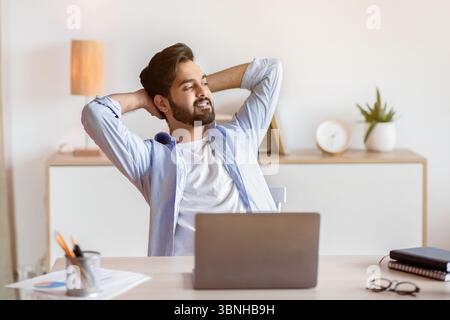 Relaxed Eastern Man Leaning Back In Chair, Resting After Work With Laptop Stock Photo