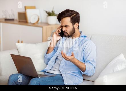 Computer Problems. Frustrated arab guy using cellphone and looking on laptop screen Stock Photo