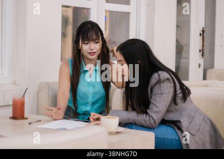 Two asian businesswomen are collaboratively reviewing project documents in a bright cafe. They are discussing strategy and innovative ideas while work Stock Photo