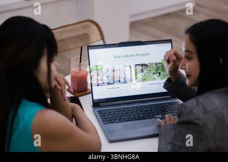 Two colleagues collaborating on a project, reviewing design concepts displayed on a laptop screen in a bright, modern workspace. Communication and tea Stock Photo
