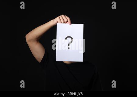 Man hiding behind paper sheet with question mark on dark background Stock Photo