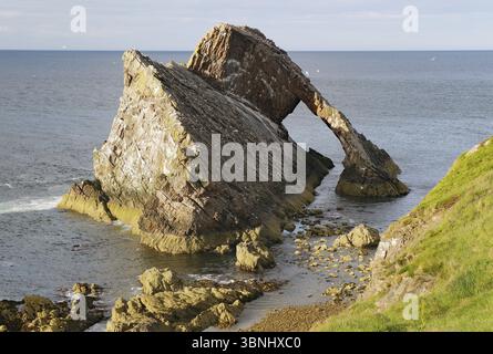 Large rock formation in a calm sea illuminated by the setting sun, Remarkable rock formation jutting into the sea under a blue sky with few clouds, Bo Stock Photo