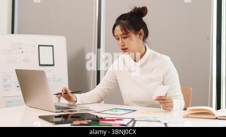 Young Asian woman user interface designer is busy working on her laptop to create new design for mobile application and web design sitting at her desk Stock Photo