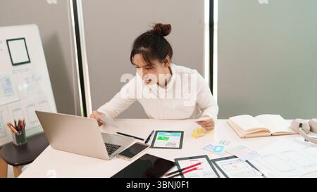 Young adult Asian woman user interface designer at her desk working on a laptop computer creating a mobile app interface. Woman designs user experienc Stock Photo
