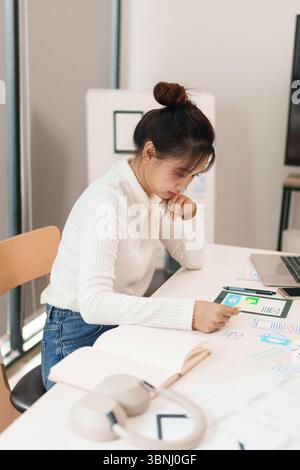 Asian adult woman designing user interface on tablet. Creative UX design process for mobile application layout with paperwork on white office desk. Stock Photo