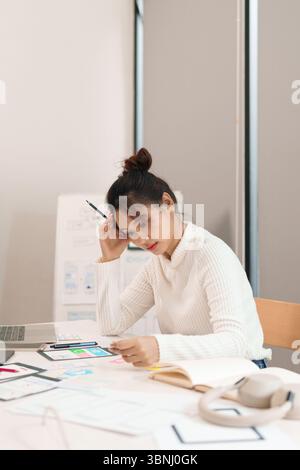 Asian adult woman UI designer is working hard on her design project in the office, creating application design with laptop and paper wireframes for a Stock Photo