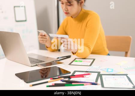 Young Asian woman UI UX designer creates mobile application user interface on a desk with laptop computer and cards. Concept for female design process Stock Photo