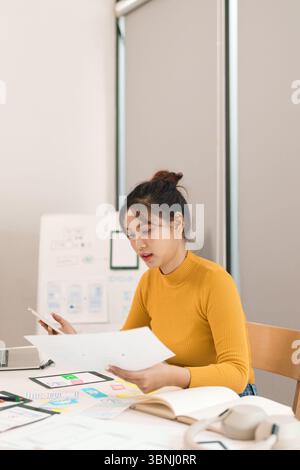 Adult Asian woman UI UX designer creating mobile app interface design. She uses mobile phone and tablet while working on mockups and wireframes in off Stock Photo