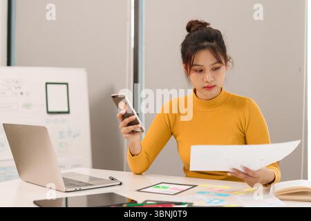An adult Asian woman, a UX UI designer, carefully examines a mobile application interface on a smartphone while reviewing paper sketches for website d Stock Photo